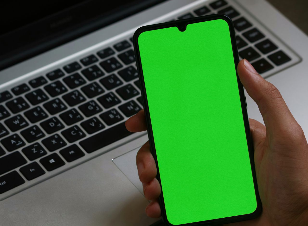 A hand holding a smartphone with a green screen over a laptop, perfect for digital mockup presentations.